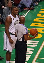 A basketball player stands on a basketball court. He wears a green jersey with the word «CELTICS» and the number 11 on the front.